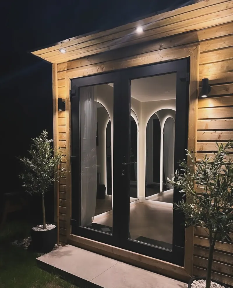A timber garden room at night, lit by soffit downlights and two outdoor wall lights, with glowing arched mirrors visible through the glass doors